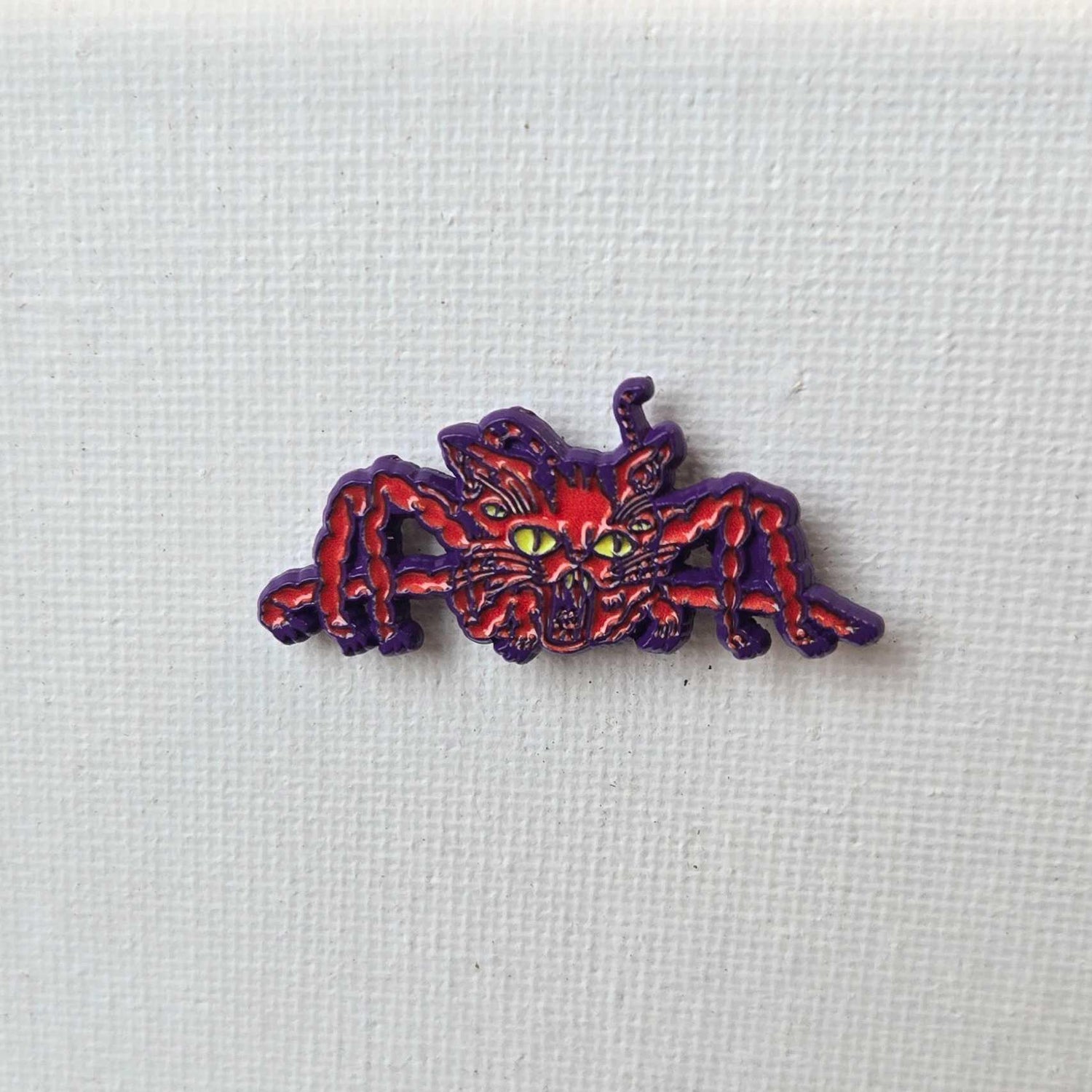 Red and purple enamel pin of a cat spider hybrid on white textured background