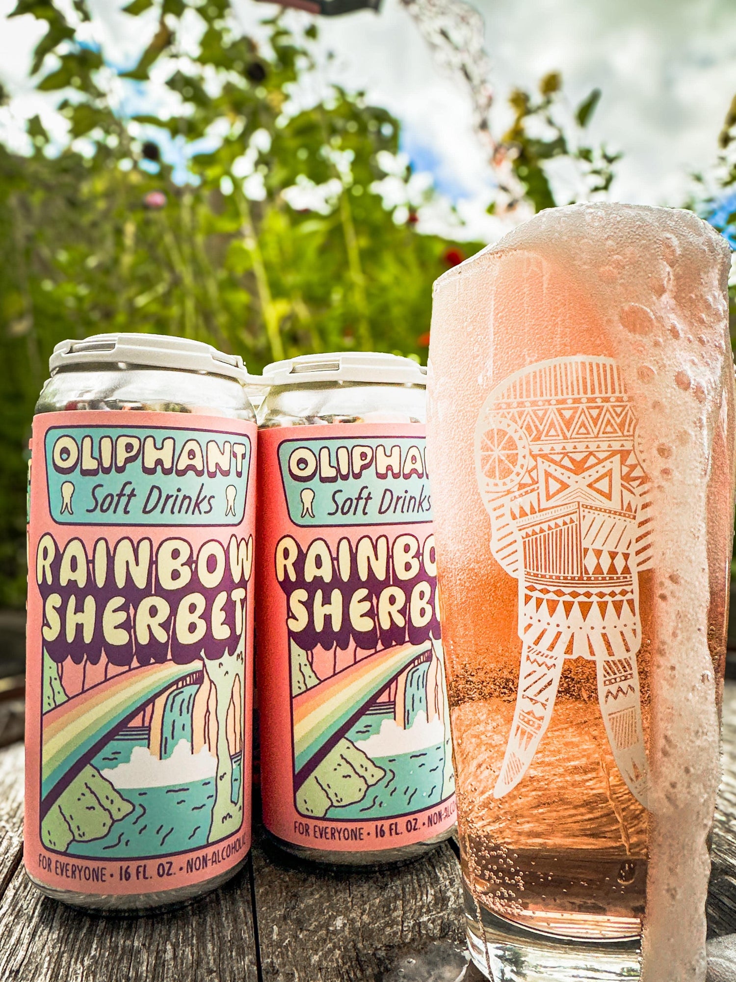 Cans of Oliphant Rainbow Sherbet soft drink next to a foamy glass on an outdoor wooden table