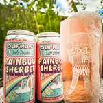 Cans of Oliphant Rainbow Sherbet soft drink next to a foamy glass on an outdoor wooden table